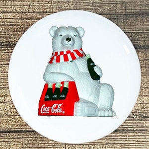 May include: A white polar bear wearing a red and white striped scarf holds a bottle of Coca-Cola and sits on a red Coca-Cola cooler with four bottles of Coca-Cola inside.