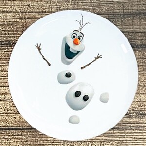 May include: A white ceramic plate with a cartoon image of Olaf, the snowman from the movie Frozen. Olaf is smiling with his arms outstretched and has a carrot nose and black eyes.