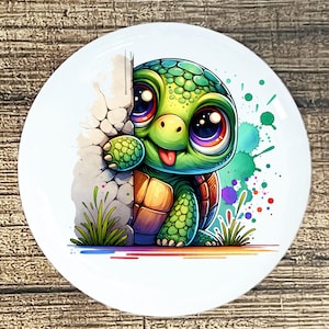 May include: A white ceramic plate featuring a cartoon turtle with large, colorful eyes and a green shell. The turtle peeks from behind a stone wall, with colorful paint splatters in the background.