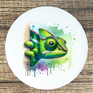 May include: A white ceramic plate featuring a vibrant illustration of a green chameleon. The chameleon is detailed with shades of green and is set against a watercolor background with splashes of blue, purple, and green.