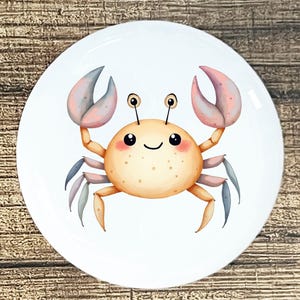May include: A white ceramic plate featuring a cartoon crab illustration. The crab has an orange body, pink and blue claws, and a smiling face. The plate is round and has a glossy finish. The crab design is centered on the plate.