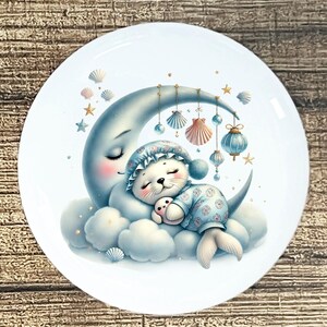 May include: A white ceramic plate featuring a whimsical illustration. A sleeping seal pup wearing a blue nightcap and pajamas rests on a crescent moon and cloud. The design includes seashells, stars, and hanging ornaments in soft pastel colors.
