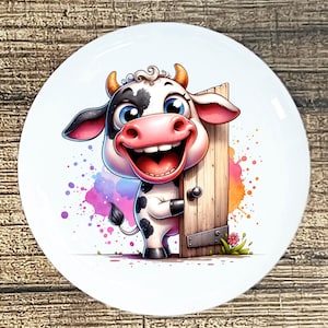 May include: A white ceramic plate featuring a cartoon cow with a big smile, black and white spots, and blue eyes. The cow is holding a wooden door, with colorful paint splatters in the background.