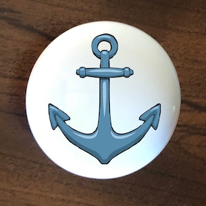 May include: A white ceramic knob with a blue anchor design. The anchor is drawn in a cartoon style with a rope loop at the top.