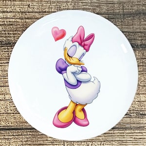 May include: White ceramic plate featuring a cartoon image of Daisy Duck. Daisy is depicted with a pink bow, purple top, and pink shoes, with a red heart above her head. The plate has a glossy finish.