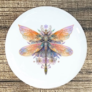 May include: White ceramic plate featuring a detailed dragonfly design. The dragonfly has colorful wings in shades of orange, purple, and gold. The design includes floral accents and intricate patterns, creating a decorative and artistic piece.