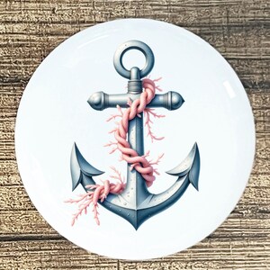 May include: A white, round decorative plate featuring a nautical anchor design. The anchor is a muted blue-gray color, wrapped with a pink rope and coral. The plate has a glossy finish and is set against a wood grain background.