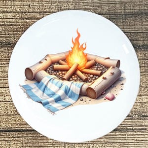 May include: White ceramic plate with a campfire scene. The design features a burning fire, logs, small stones, a blue and white striped blanket, and a seashell. The plate has a glossy finish.