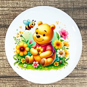 May include: A white ceramic plate featuring a cartoon illustration of a yellow bear wearing a red shirt, surrounded by colorful flowers and a friendly bee. The bear is holding a yellow flower, creating a cheerful and whimsical design.