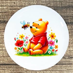 May include: A white ceramic plate featuring a colorful illustration of a smiling yellow bear in a red shirt, sitting in a field of flowers. A cartoon bee hovers near the bear's face. The plate has a glossy finish.