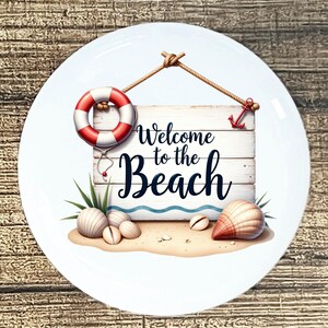 May include: White ceramic plate with a beach-themed design. Features a wooden sign with the text "Welcome to the Beach", a red and white life preserver, seashells, and an anchor. The plate has a sandy beach scene.