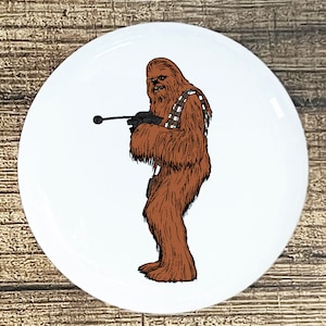 May include: A white button with a black and white line drawing of Chewbacca from Star Wars. Chewbacca is holding a blaster rifle and is wearing a bandolier.