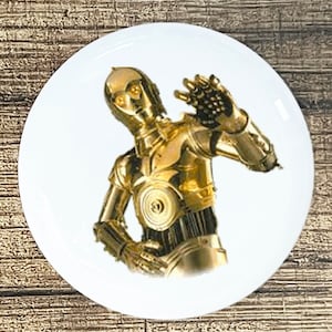 May include: A gold robot with a human-like face and a single eye on each side of its head. The robot has a raised arm with a mechanical hand. The robot is standing on a white background.
