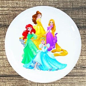 May include: A white round sticker with a colorful image of five Disney princesses. The princesses are wearing their signature gowns and have long flowing hair. The princesses are Cinderella, Belle, Ariel, Rapunzel, and Snow White.