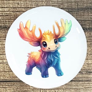 May include: A white ceramic plate featuring a cartoon moose with rainbow-colored antlers and fur. The moose has large, expressive eyes and a friendly smile. The color palette includes shades of orange, yellow, blue, and purple.
