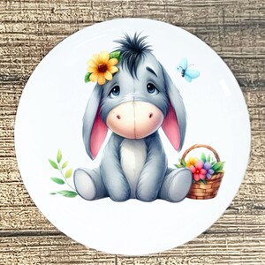 May include: A white ceramic plate featuring a watercolor illustration of a gray donkey with a yellow flower crown. A blue butterfly and a basket of flowers are also depicted. The donkey is sitting down.