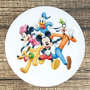 May include: White ceramic plate featuring a colorful illustration of Disney characters Mickey Mouse, Minnie Mouse, Donald Duck, Goofy, and Pluto. The characters are depicted in a running pose, with bright colors and detailed artwork.