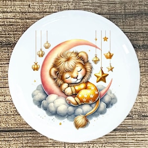 May include: A white ceramic plate featuring a whimsical illustration of a sleeping lion cub. The lion rests on a crescent moon and cloud, surrounded by hanging gold stars and lanterns. The cub wears a yellow outfit with crown patterns.