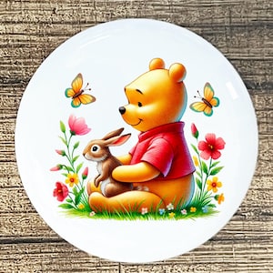 May include: A white ceramic plate featuring a cartoon illustration of a yellow bear in a red shirt hugging a brown rabbit. Colorful flowers and butterflies surround the characters, set against a green grassy background.