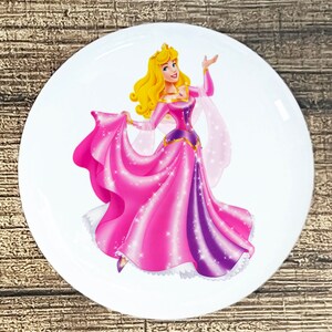 May include: A white round plate with a cartoon image of a princess wearing a pink and purple dress with a crown. The princess has long blonde hair and is smiling.