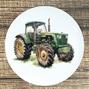 May include: A white ceramic plate featuring a watercolor painting of a green John Deere tractor. The tractor has large tires and is set against a light green and white background. The plate is round and has a glossy finish.