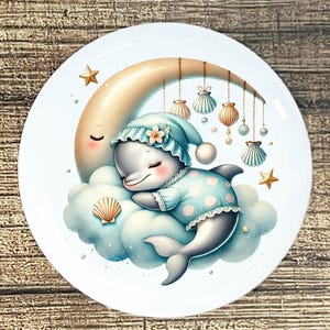 May include: A white ceramic plate featuring a whimsical illustration of a sleeping dolphin. The dolphin, wearing a blue nightcap and pajamas, rests on a cloud beneath a crescent moon. Decorative seashells and stars adorn the design, creating a dreamy, nautical theme.