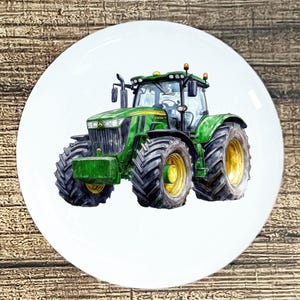 May include: A white ceramic plate featuring a detailed illustration of a green and yellow tractor. The tractor has large black tires and is set against a white background. The plate is round and appears to be new.