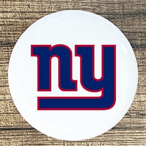 May include: A white round button with the New York Giants logo. The logo is blue with a red outline and the letters "NY" in a bold font.