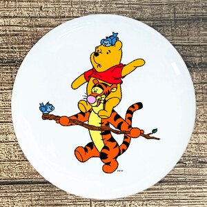 May include: A white ceramic round button with a cartoon image of Winnie the Pooh and Tigger. Winnie the Pooh is sitting on Tigger's back, and Tigger is holding a stick with a blue bird on it.  The background is white.