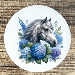 May include: A white ceramic plate featuring a detailed illustration of a horse's head with a gray and white coat. The horse is surrounded by blue hydrangea flowers and green leaves, creating a floral design.