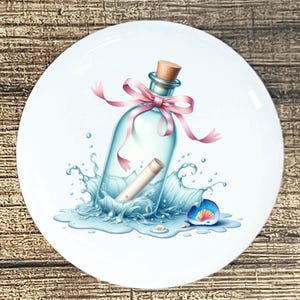 May include: A white ceramic plate featuring a whimsical illustration of a message in a bottle. The clear glass bottle is adorned with a pink ribbon and sits amidst a splash of water. A seashell is also present.