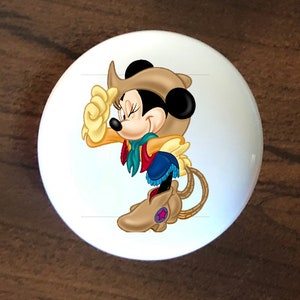 May include: A white ceramic knob with a cartoon image of Minnie Mouse dressed as a cowgirl. She is wearing a brown cowboy hat, a red bandana, a blue denim skirt, and brown cowboy boots. She is tipping her hat and smiling.