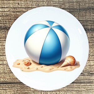 May include: A white ceramic plate featuring a beach scene. A blue and white beach ball sits on a sandy surface with two seashells. The plate has a glossy finish and is set against a wooden background.
