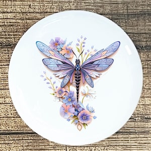 May include: A white ceramic plate featuring a detailed illustration of a dragonfly with lavender and blue wings. The dragonfly is surrounded by delicate purple and peach flowers and foliage. The plate has a glossy finish.