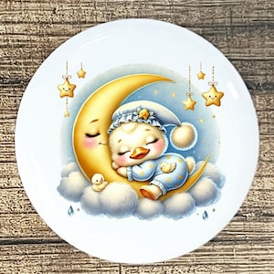 May include: A white ceramic plate featuring a whimsical illustration of a sleeping duckling wearing a nightcap, nestled on a crescent moon. The duckling is surrounded by clouds, stars, and a small duckling. The design has a soft, pastel color palette.