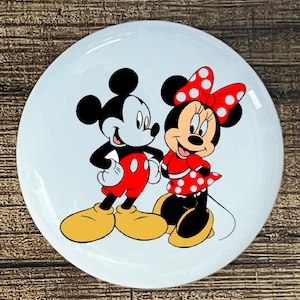 May include: A white ceramic plate with a cartoon image of Mickey Mouse and Minnie Mouse. Mickey is wearing red shorts and yellow shoes. Minnie is wearing a red dress with white polka dots and a red bow with white polka dots.