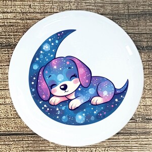 May include: A white, round ceramic plate featuring a cartoon puppy sleeping on a crescent moon. The puppy and moon are decorated with a galaxy design in shades of blue, purple, and white. The puppy has closed eyes and a smiling expression.