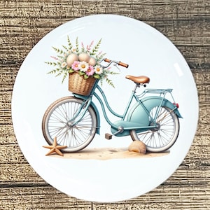 May include: A decorative white plate featuring a light blue bicycle with a wicker basket filled with pink and white flowers. A starfish and seashell are on the sand. The bicycle has a brown seat and handlebars.