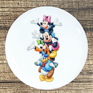 May include: White ceramic plate featuring a stack of Disney characters: Minnie Mouse, Mickey Mouse, Goofy, and Donald Duck. The characters are in full color, with Minnie wearing a pink bow, Mickey in blue, Goofy in orange, and Donald in blue.