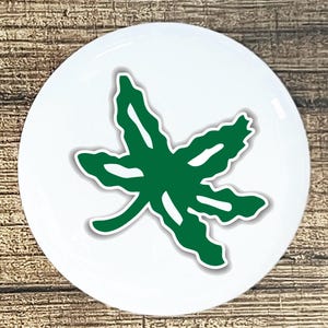 May include: A white, round decorative item featuring a green leaf design with a white outline. The leaf is centered on the white surface. The item is set against a wooden background.