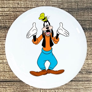 May include: A white round button with a cartoon image of Goofy. Goofy is wearing a yellow hat, an orange shirt, a black vest, blue pants, and brown shoes. He is smiling and has his arms outstretched.