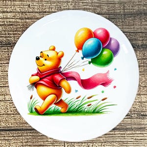 May include: A white, round plate featuring a cartoon illustration of a yellow bear wearing a red scarf and shirt, holding colorful balloons. The bear is walking through green grass with brown reeds.