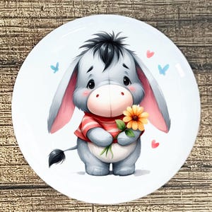 May include: A white ceramic plate featuring a cartoon donkey character. The donkey is gray with a red and white striped shirt and is holding a yellow flower. Pink hearts and blue butterflies decorate the plate.