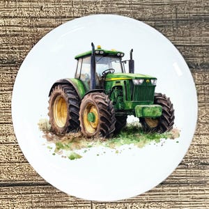 May include: White ceramic plate featuring a watercolor illustration of a green tractor. The tractor has large black tires with yellow rims and is set against a backdrop of grass and dirt. The plate is round and has a glossy finish.