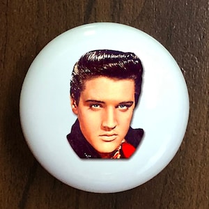 May include: A white ceramic knob with a color image of Elvis Presley. The image shows Elvis with his signature pompadour hairstyle and a serious expression. He is wearing a black jacket and a red scarf.