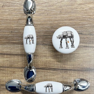 May include: Three ceramic cabinet knobs featuring a white background with a black and white illustration of an AT-AT walker from Star Wars. The knobs are in the shape of a round knob and two oval knobs with a silver metal base.