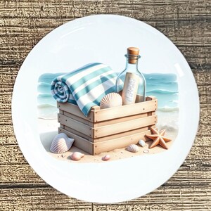 May include: A decorative plate features a beach scene with a wooden crate holding a rolled blue and white striped towel, seashells, and a bottle with a message. A starfish and more shells are scattered on the sand.