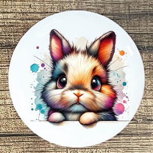 May include: A white ceramic plate featuring a colorful illustration of a bunny with large, expressive eyes. The bunny's fur is a mix of orange, pink, blue, and white, with colorful paint splatters around it. The plate is round and has a glossy finish.