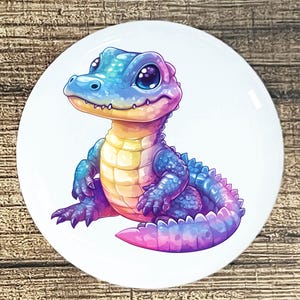 May include: A round, white ceramic plate featuring a colorful illustration of a cartoon alligator. The alligator is depicted in shades of blue, purple, pink, and yellow, with large, expressive eyes. The design is whimsical and vibrant, suitable for home decor.