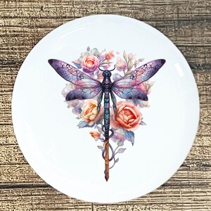 May include: White ceramic plate featuring a watercolor illustration of a dragonfly with purple and blue wings, set against a backdrop of pink and peach roses and foliage. The dragonfly's body is a gradient of blues and greens.